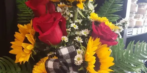 A floral arrangement featuring bright yellow sunflowers and red roses accented with green ferns and a black-and-white plaid ribbon.