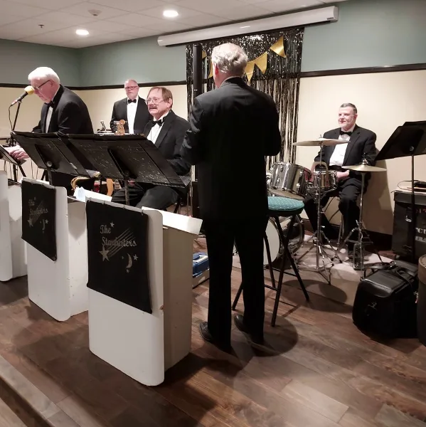 A five-piece band performing on a stage with drums, keyboard, and brass instruments.