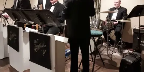 A five-piece band performing on a stage with drums, keyboard, and brass instruments.