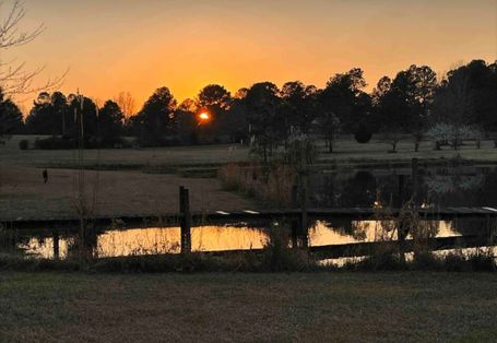 Countryside property at sunset with open fields, trees, and a lake in the foreground.