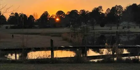 Countryside property at sunset with open fields, trees, and a lake in the foreground.