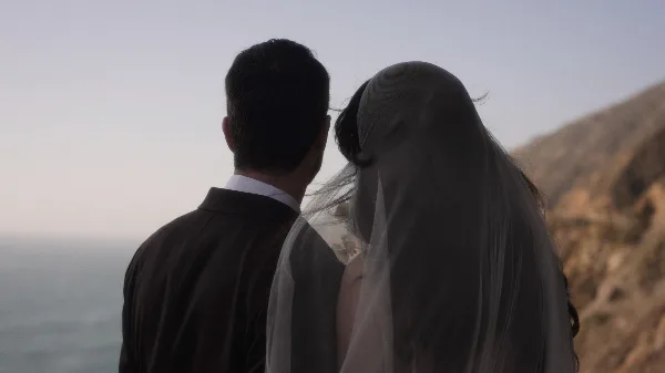 A cinematic shot of a bride and groom standing together, viewed from behind, looking out at a coastal landscape.