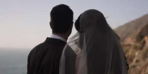 A cinematic shot of a bride and groom standing together, viewed from behind, looking out at a coastal landscape.