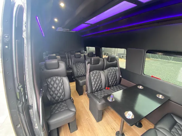 Interior view of a luxury passenger van with black quilted leather seats, a table, and purple ambient ceiling lighting.
