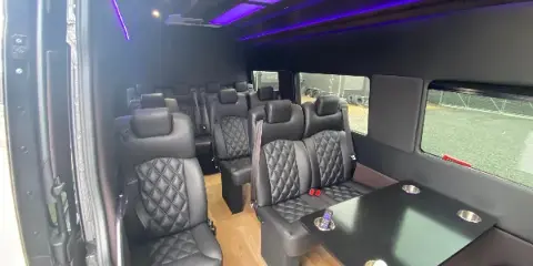 Interior view of a luxury passenger van with black quilted leather seats, a table, and purple ambient ceiling lighting.