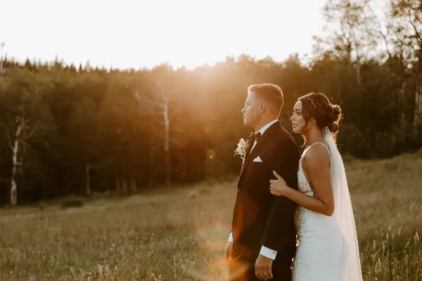 A bride and groom standing in a grassy field at sunset, with the bride resting her hand on the groom's arm.
