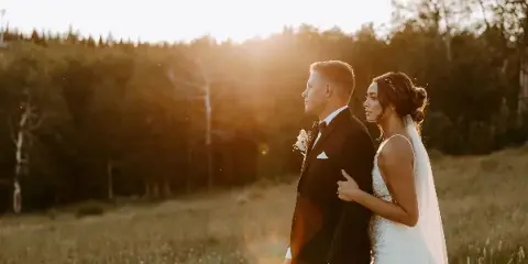 A bride and groom standing in a grassy field at sunset, with the bride resting her hand on the groom's arm.