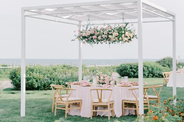 An outdoor wedding table setting under a white pergola with a large hanging floral arrangement and ocean view in the background.