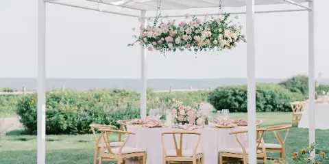 An outdoor wedding table setting under a white pergola with a large hanging floral arrangement and ocean view in the background.