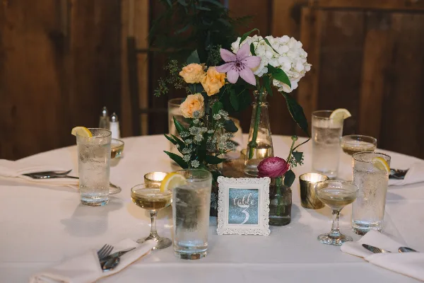 A wedding reception table setting featuring a floral centerpiece with white and peach flowers, water glasses, and a table number sign.