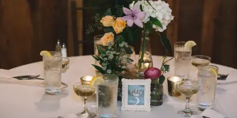A wedding reception table setting featuring a floral centerpiece with white and peach flowers, water glasses, and a table number sign.