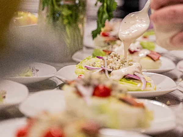 A hand pouring creamy dressing over a freshly prepared salad with lettuce, bacon, and red onion on a white plate.