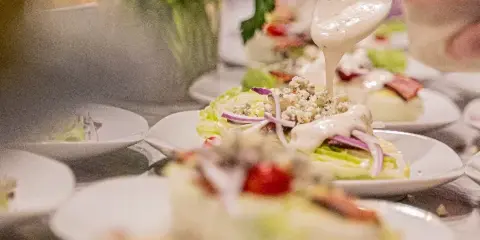A hand pouring creamy dressing over a freshly prepared salad with lettuce, bacon, and red onion on a white plate.