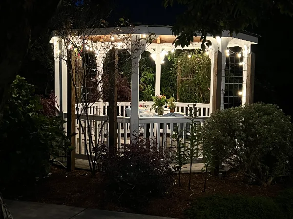 A white wooden gazebo illuminated by string lights at night, featuring floral arrangements and surrounded by garden greenery.