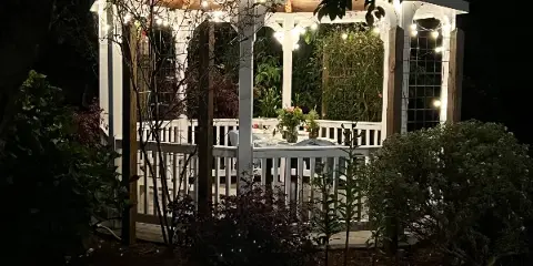 A white wooden gazebo illuminated by string lights at night, featuring floral arrangements and surrounded by garden greenery.