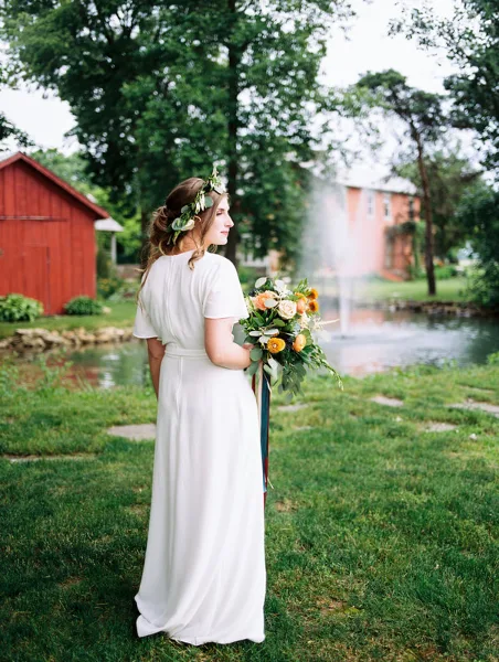 A bride in a white dress stands on the grass holding a bouquet, with a pond and red barn in the background.