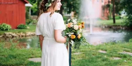 A bride in a white dress stands on the grass holding a bouquet, with a pond and red barn in the background.