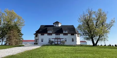 Exterior view of a white historic barn wedding venue with a dark roof and cupola on a grassy lawn under a blue sky.