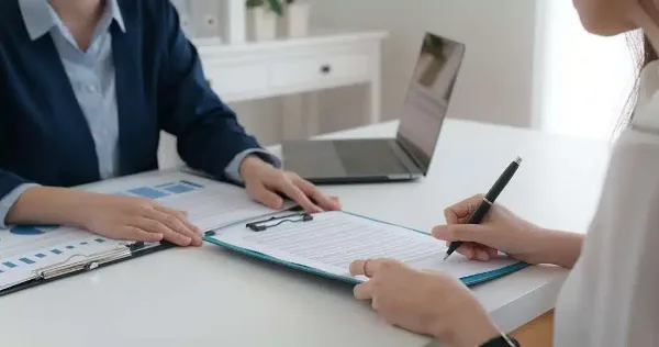 A professional in a business suit and a client signing a document on a clipboard at a desk.