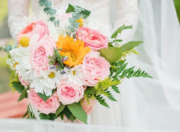 A wedding bouquet of pink garden roses, white daisies, a yellow sunflower, and greenery held in front of a white wedding dress.