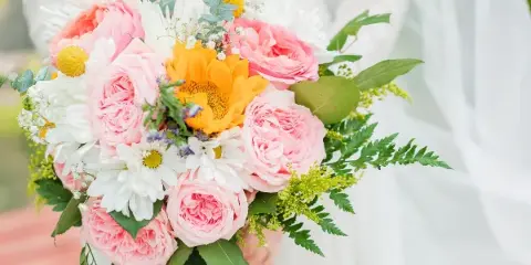 A wedding bouquet of pink garden roses, white daisies, a yellow sunflower, and greenery held in front of a white wedding dress.