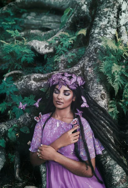 A woman with long dark hair and a purple butterfly headpiece poses outdoors against a backdrop of tree roots and greenery, wearing a purple floral dress.