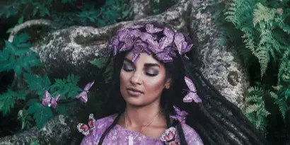 A woman with long dark hair and a purple butterfly headpiece poses outdoors against a backdrop of tree roots and greenery, wearing a purple floral dress.