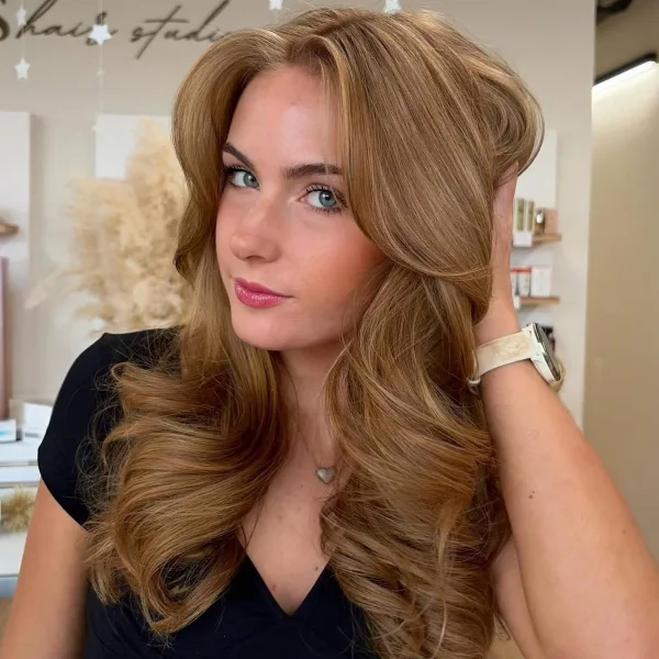 A woman with long, styled blonde hair and makeup posing in a salon with the Bliss Hair Studio logo visible on the wall behind her.