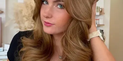 A woman with long, styled blonde hair and makeup posing in a salon with the Bliss Hair Studio logo visible on the wall behind her.