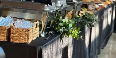 A catering buffet line featuring stainless steel chafing dishes, serving baskets, and a black tablecloth at an event venue.