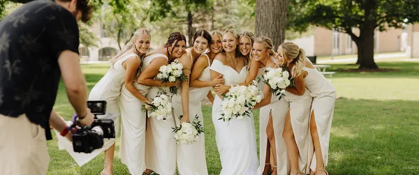A videographer filming a bride and her bridesmaids holding white bouquets outdoors.