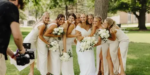 A videographer filming a bride and her bridesmaids holding white bouquets outdoors.