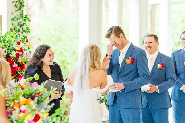 A wedding officiant in a black dress stands at an altar with a bride and groom during a ceremony, with floral arrangements in the foreground.