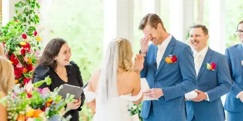 A wedding officiant in a black dress stands at an altar with a bride and groom during a ceremony, with floral arrangements in the foreground.