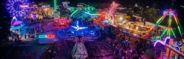 Aerial view of the Colusa County Fairgrounds at night, illuminated with colorful neon lights and featuring a large Ferris wheel and other amusement rides.