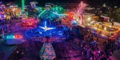 Aerial view of the Colusa County Fairgrounds at night, illuminated with colorful neon lights and featuring a large Ferris wheel and other amusement rides.