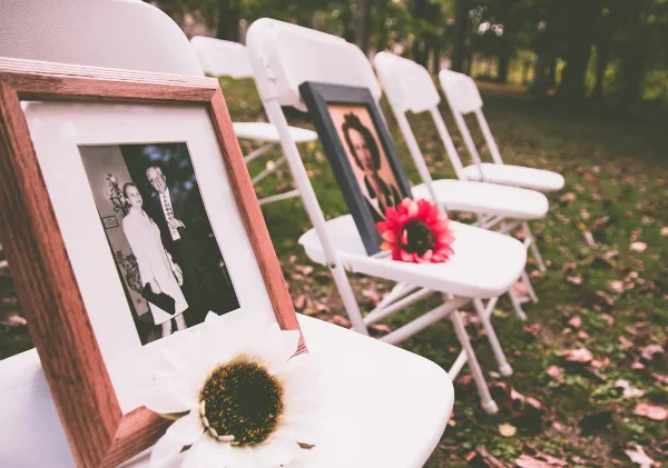 White folding chairs arranged in a row outdoors with framed photos and flowers placed on the seats.
