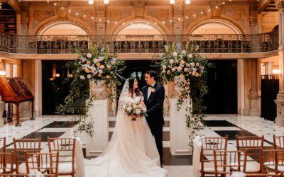 A bride and groom standing at the altar of a grand, ornate venue with large floral arrangements and styled seating.