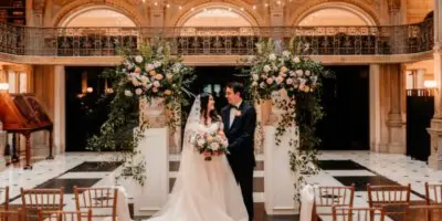 A bride and groom standing at the altar of a grand, ornate venue with large floral arrangements and styled seating.