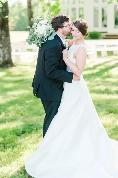 A bride and groom embracing in a grassy outdoor area with a white building in the background.