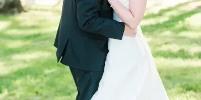 A bride and groom embracing in a grassy outdoor area with a white building in the background.