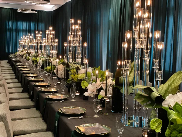 A long wedding reception table decorated with white orchids, greenery, and tall glass candle holders against a dark teal curtain backdrop.