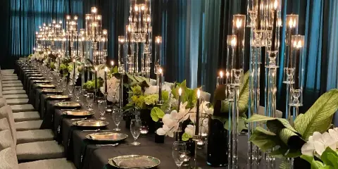 A long wedding reception table decorated with white orchids, greenery, and tall glass candle holders against a dark teal curtain backdrop.