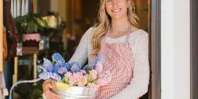 A smiling florist in a striped apron holds a metal bucket filled with blue and pink hyacinths at the entrance of a flower shop.