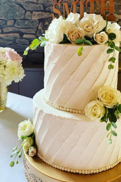 A two-tier white wedding cake with diagonal frosting texture, decorated with white and cream roses and greenery, featuring a wooden 'SMITH' topper.