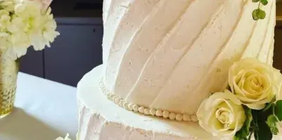 A two-tier white wedding cake with diagonal frosting texture, decorated with white and cream roses and greenery, featuring a wooden 'SMITH' topper.