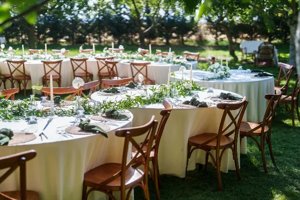 An outdoor wedding reception setup featuring round tables with white linens, greenery centerpieces, and wooden chairs on a lawn.