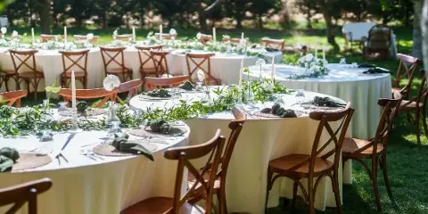 An outdoor wedding reception setup featuring round tables with white linens, greenery centerpieces, and wooden chairs on a lawn.