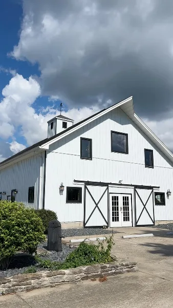 Exterior view of a white barn wedding venue with black trim and a cupola under a cloudy sky.