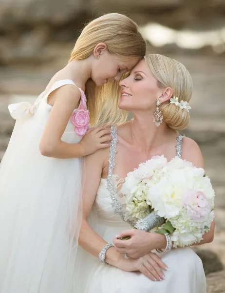 A bride with a styled blonde updo and floral hair accessory holding a white bouquet, embracing a young girl in a white dress.